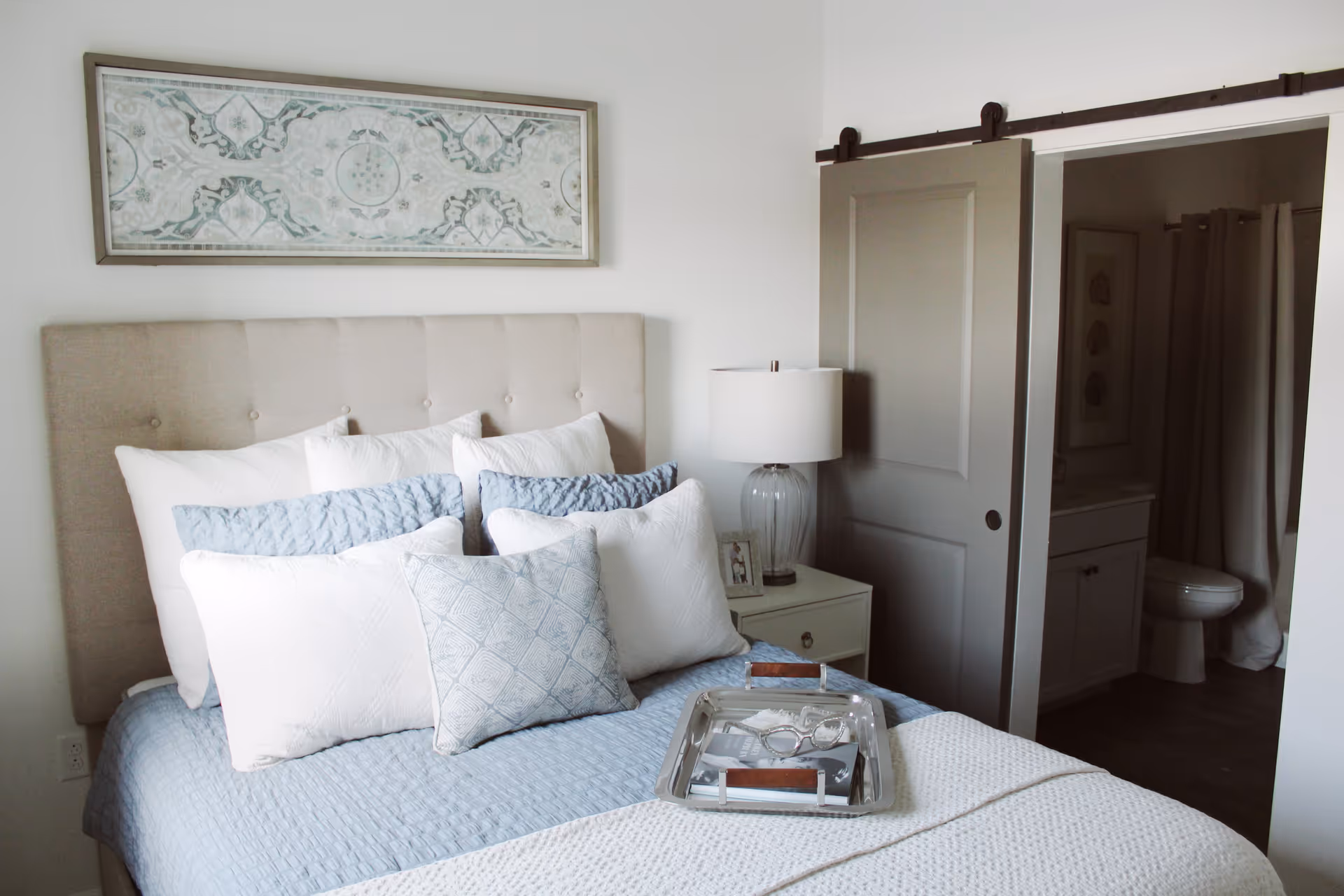 A neatly made bedroom with a tufted headboard, layered pillows, a bedside table and lamp, and a sliding barn door opening to a bathroom.