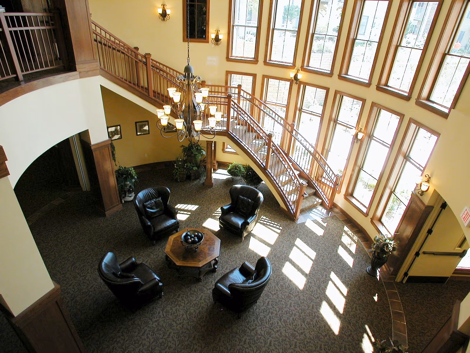 A spacious, well-lit common area with large windows allowing natural light to flood in. The room features a circular arrangement of four black leather armchairs around a wooden octagonal coffee table. A grand chandelier hangs from the ceiling above the seating area. A staircase with wooden railings curves upward along the wall of windows. The walls are painted a warm yellow, and there are several wall sconces providing additional lighting. Potted plants and framed pictures decorate the space.