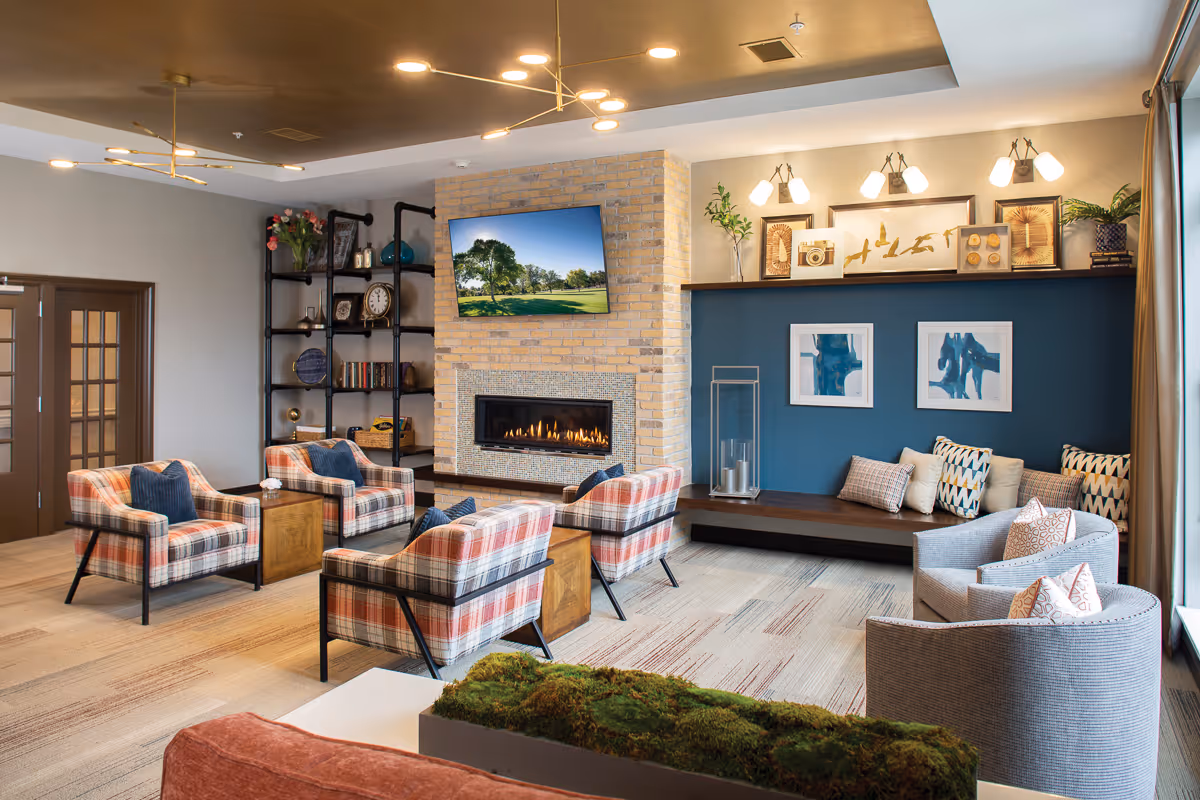 A cozy living room area in St. Rita Square featuring a modern fireplace with a TV mounted above it, surrounded by four plaid armchairs and two gray armchairs. The room has a blue accent wall with framed artwork and a built-in bench with cushions. There is a black metal shelving unit with decorative items and a wooden floor with a patterned carpet.