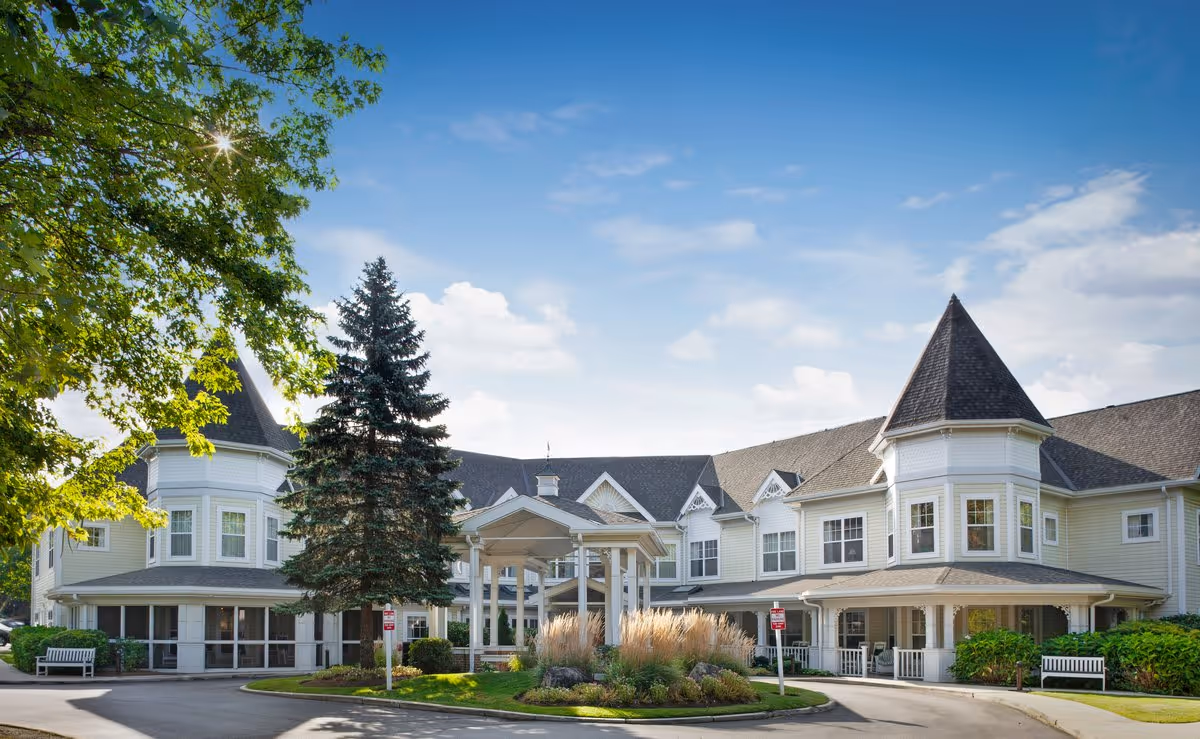 Front entrance of a large two-story senior living building with turrets, a covered porte-cochère, driveway and landscaped grounds under a blue sky.