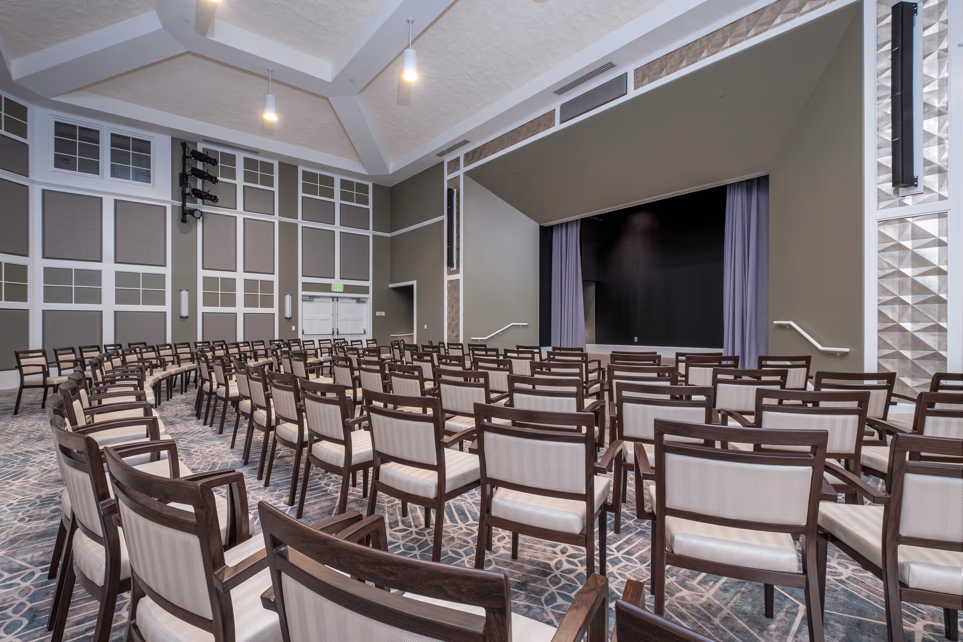 An empty auditorium with rows of wooden chairs facing a small stage with curtains. The room has high ceilings with hanging lights and decorative wall panels.