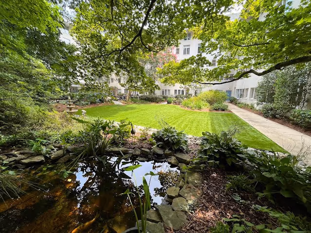 A serene outdoor garden area at Saint Anne's Terrace featuring a small pond with surrounding rocks and plants, a well-maintained green lawn, various shrubs and trees, and a paved walkway leading through the garden with a multi-story building in the background.