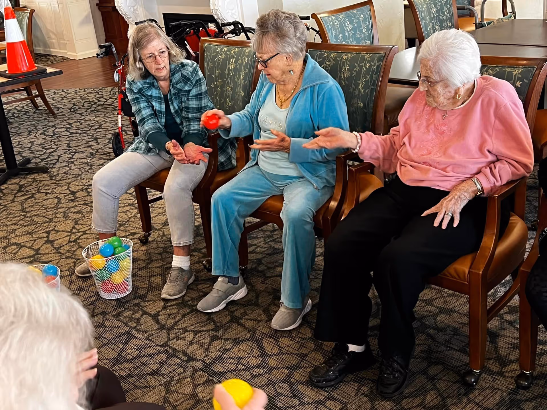 Three elderly women seated in a senior living common room passing colorful plastic balls during an activity.