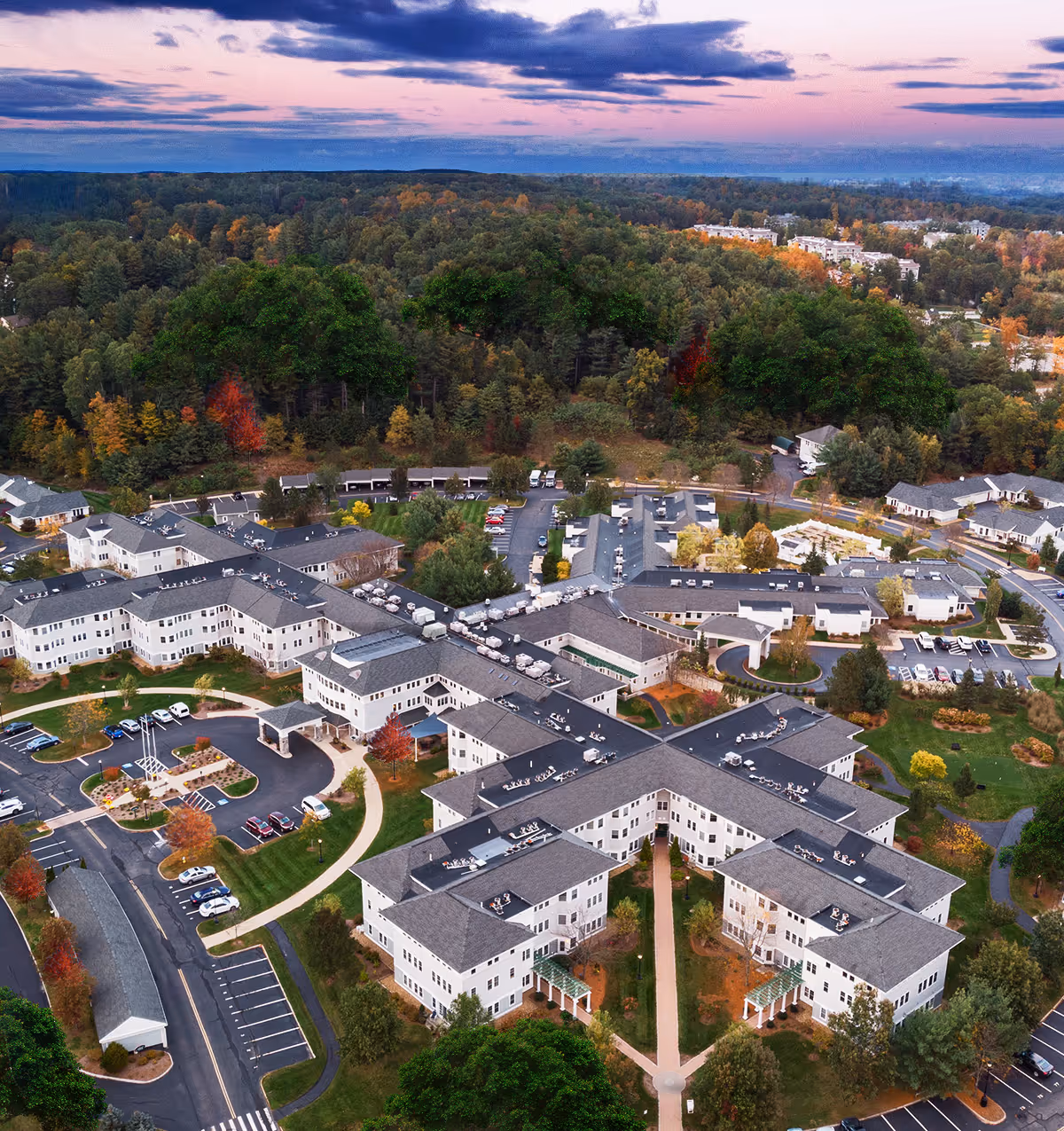 Aerial view of The Huntington At Nashua senior living facility surrounded by trees and greenery, with multiple connected buildings, parking lots, and pathways under a colorful sky at sunset.