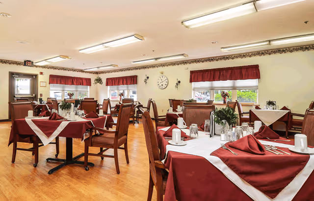 A dining room in a senior living facility with multiple tables covered in burgundy and white tablecloths, set with cups, glasses, silverware, and napkins. The room has wooden chairs, large windows with red valances, a clock on the wall, and a wooden floor.