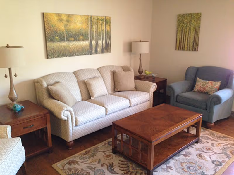 A cozy living room with a light-colored patterned sofa, a blue armchair with a floral pillow, two wooden side tables each with a lamp, a wooden coffee table, and two paintings of trees on the walls.