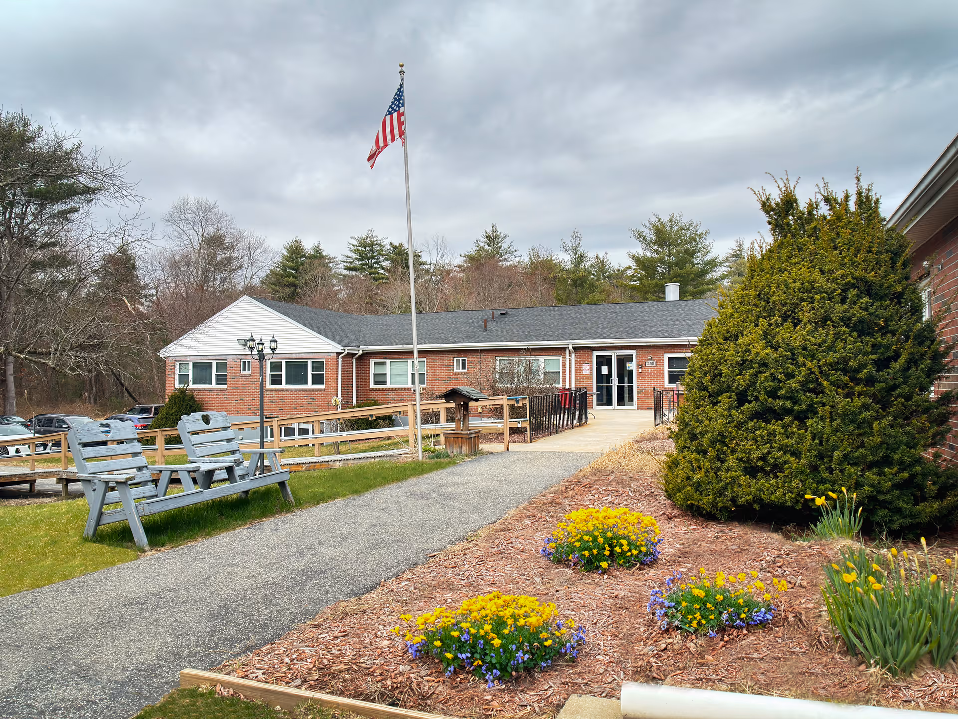 Exterior view of Palmer Healthcare Center showing a single-story brick building with a paved walkway leading to the entrance. There is an American flag on a flagpole in front of the building, benches on the left side, and landscaped flower beds with yellow and purple flowers on the right. Trees and parked cars are visible in the background under a cloudy sky.