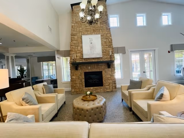 Bright common living room with beige armchairs arranged around a tufted ottoman facing a tall stone fireplace beneath a chandelier.