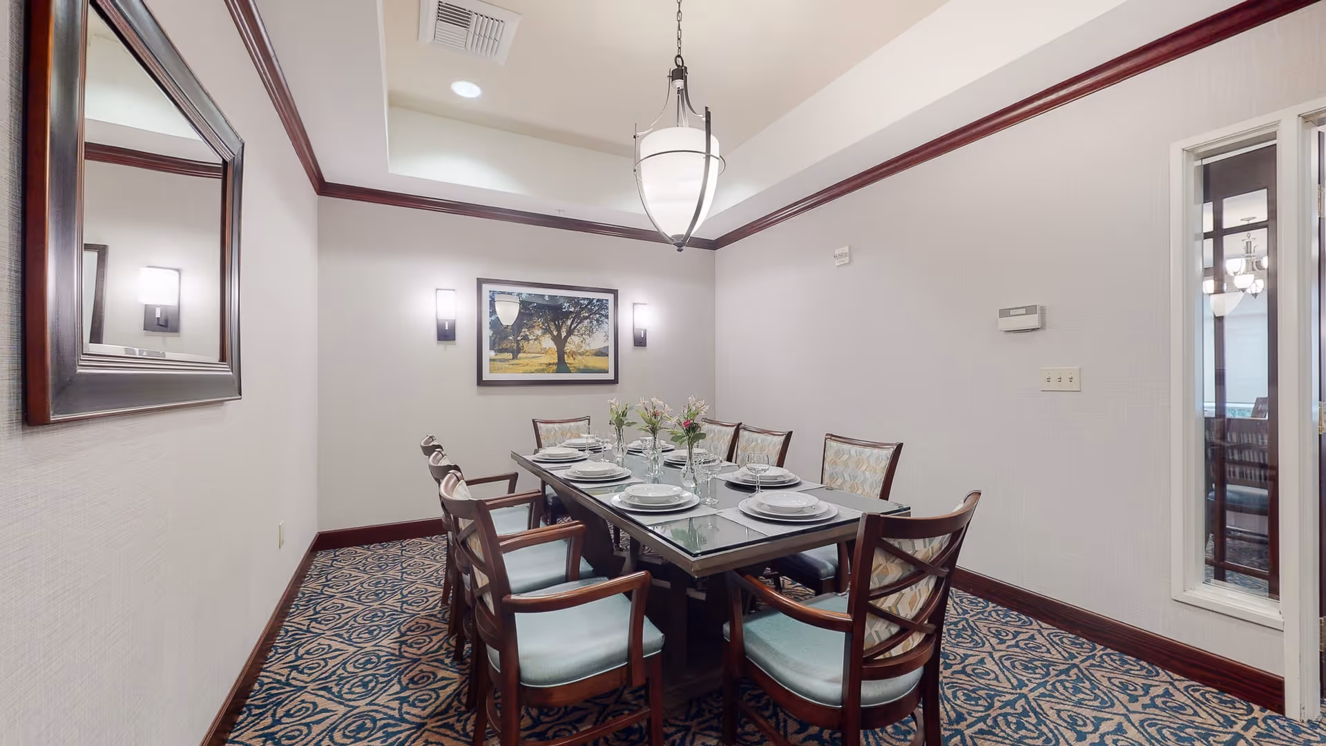 A formal dining room with a long rectangular table set for eight people. The table has place settings with plates, glasses, and napkins, and small flower arrangements as centerpieces. The room features patterned carpet, light-colored walls with dark wood trim, a large mirror on one wall, a framed picture of a tree on another wall, and a hanging light fixture above the table.