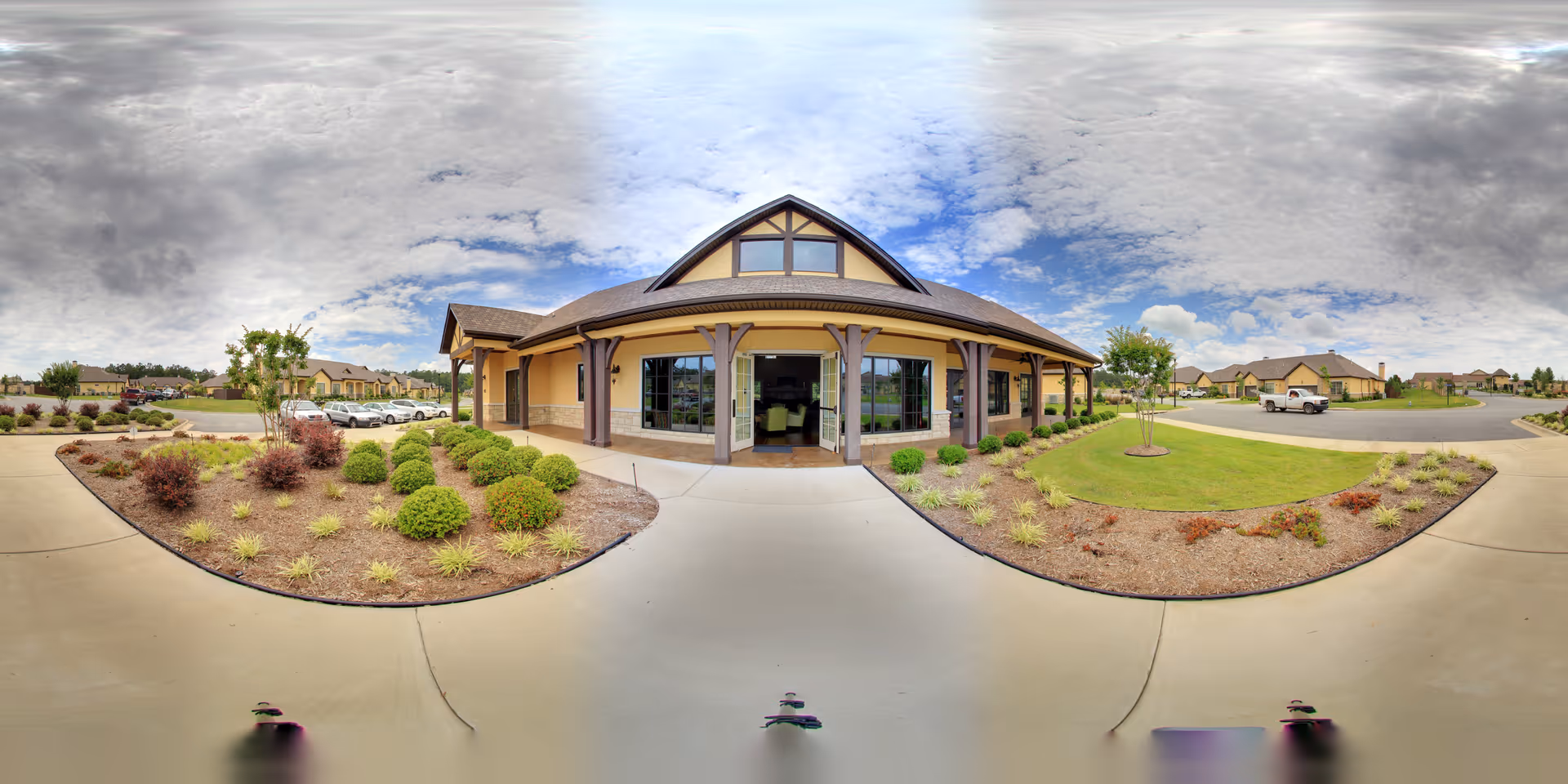 Front entrance of a single-story senior living building with a covered porch, landscaped beds, and a circular driveway under a cloudy sky.