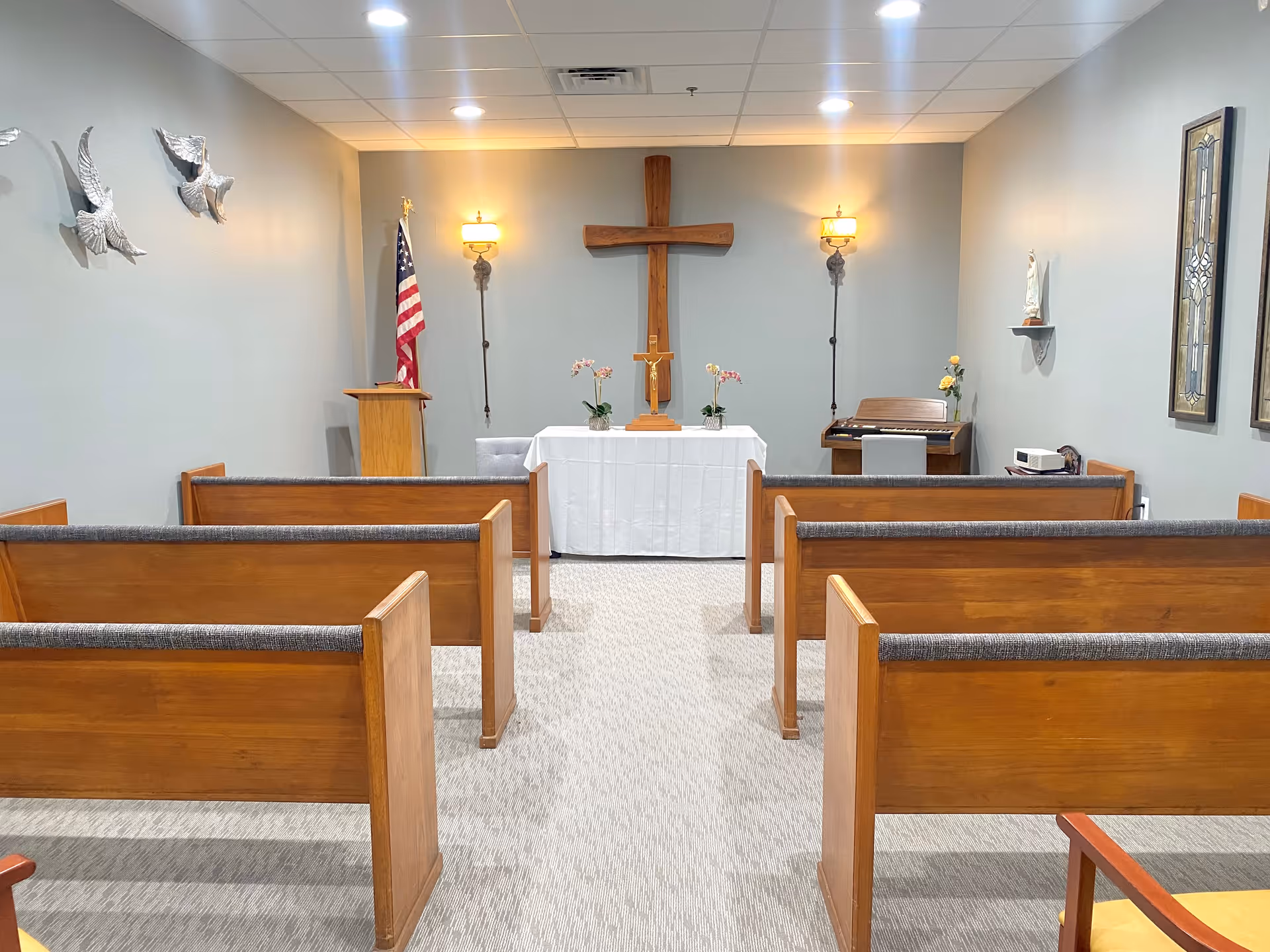 Interior view of a small chapel room with wooden pews arranged in rows facing a white altar table. Behind the altar is a large wooden cross mounted on the wall, flanked by two wall sconces with lights. An American flag stands to the left of the altar, and a small organ with a vase of flowers is on the right. The walls are light gray with decorative elements including metal bird sculptures and framed stained glass panels.