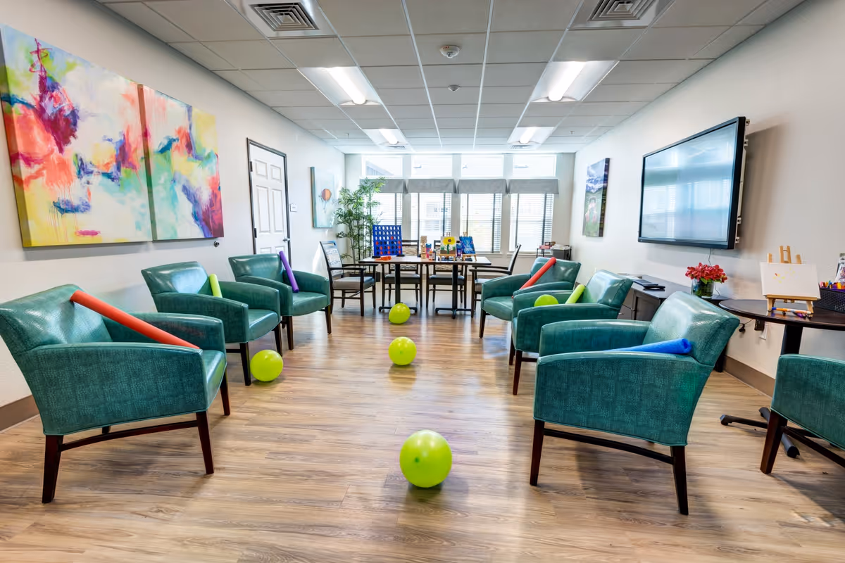 A bright activity room with six teal armchairs arranged in a semi-circle, each with a colorful foam noodle on the seat. Green balloons are scattered on the wooden floor. At the back, there is a table with chairs and board games, and large windows with blinds letting in natural light. The walls are decorated with colorful abstract paintings and a large flat-screen TV is mounted on the right wall.