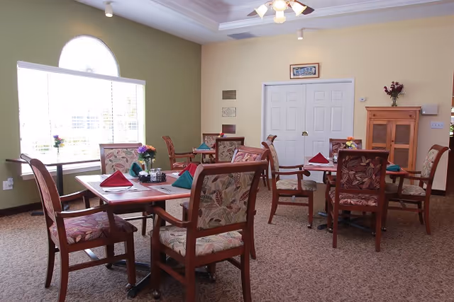A dining room with several wooden tables and chairs arranged neatly. Each table has folded cloth napkins in red and green, small flower vases, and condiment holders. The room has a large arched window letting in natural light, light green and beige walls, a ceiling fan with lights, and a wooden cabinet against one wall.