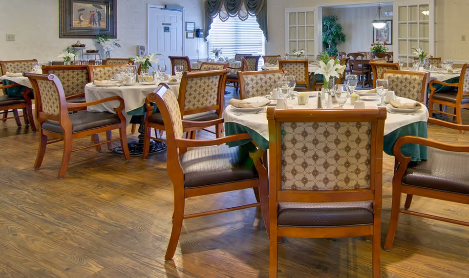 A dining room in a senior living facility with multiple round tables covered with white tablecloths and green skirts. Each table is set with glassware, napkins, and floral centerpieces. Wooden chairs with patterned upholstery surround the tables. The room has wooden flooring, a window with draped curtains, and a framed painting on the wall.