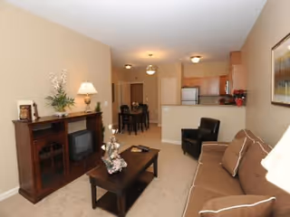 Interior view of a living room and kitchen area in an active adult retirement community. The living room features a brown sofa with cushions, a dark wood coffee table with a floral centerpiece, a matching entertainment cabinet with a lamp and decorative items, and a black armchair. The kitchen area includes wooden cabinets, a refrigerator, and a dining table with chairs. The walls are painted beige and the floor is carpeted.