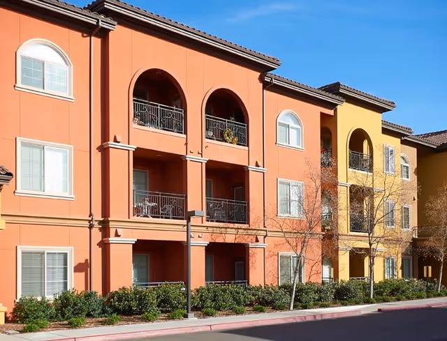 Exterior front of a three-story terracotta and yellow apartment building with arched balconies, railings, and landscaped shrubs.