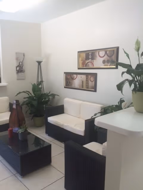 A bright and clean living room area with white walls and tiled floor. The room features black wicker furniture with white cushions, including a loveseat and chairs around a black glass coffee table. There are several green potted plants placed around the room, a tall floor lamp in the corner, and two abstract framed artworks hanging on the wall.