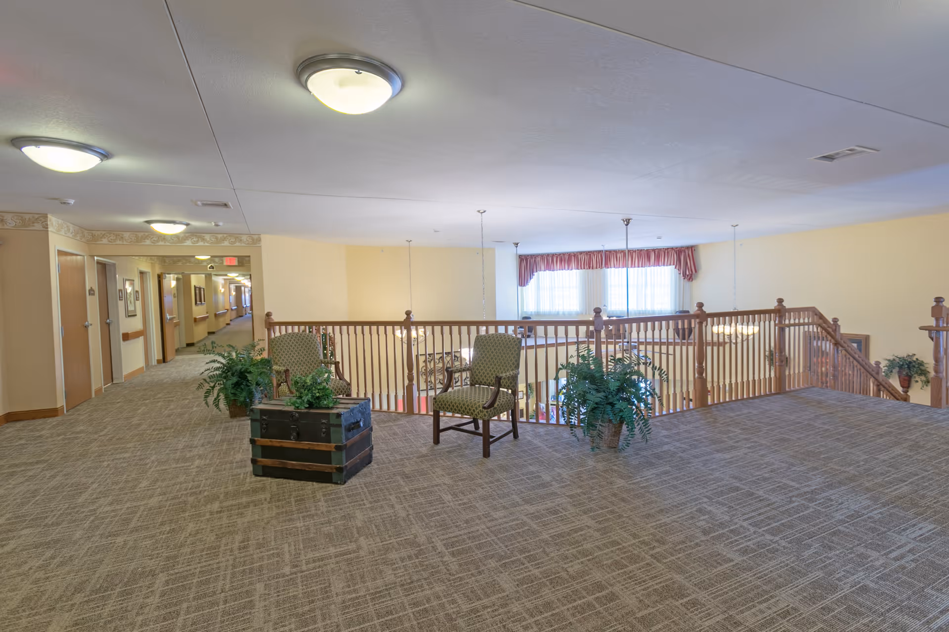 A spacious indoor hallway area in a senior living facility with beige carpet and walls. There are two green upholstered chairs and a decorative trunk placed near a wooden railing overlooking a lower level. Several potted plants are arranged around the seating area. The hallway extends to the left with multiple doors and framed pictures on the walls. Ceiling lights illuminate the space.