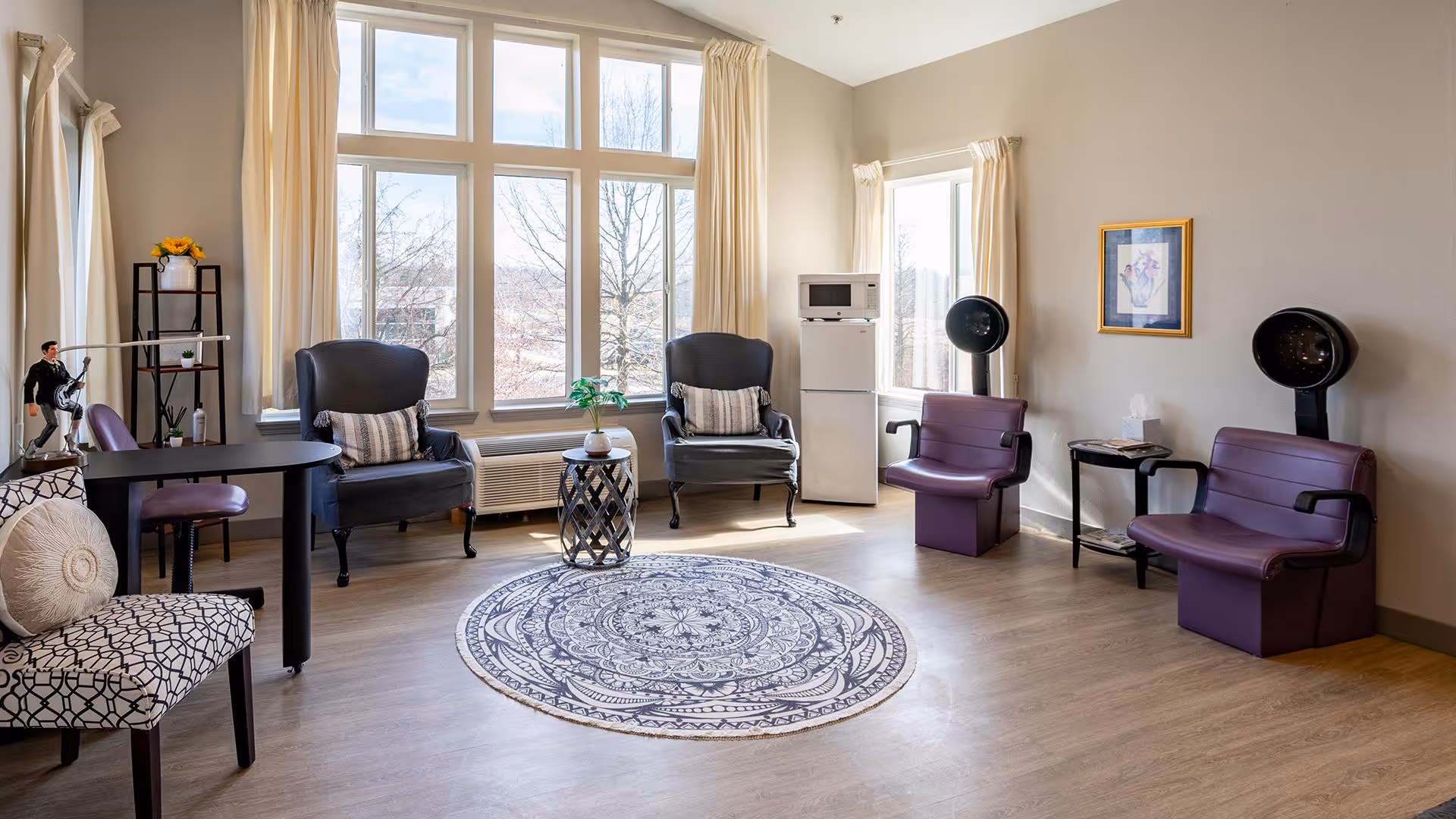 A bright and spacious room with large windows letting in natural light. The room features a round patterned rug in the center, two black armchairs with striped cushions near the windows, two purple salon chairs with hair dryers, a small table with a tissue box, a mini fridge with a microwave on top, and a decorative shelf with a figurine and a vase with yellow flowers.