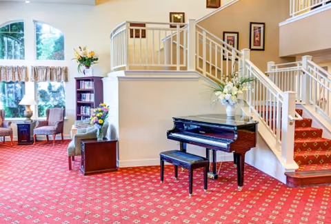 A bright and spacious living area with a red patterned carpet, a black grand piano with a matching bench, floral arrangements on the piano and side tables, comfortable chairs near large windows with curtains, and a staircase with white railings leading to an upper level.