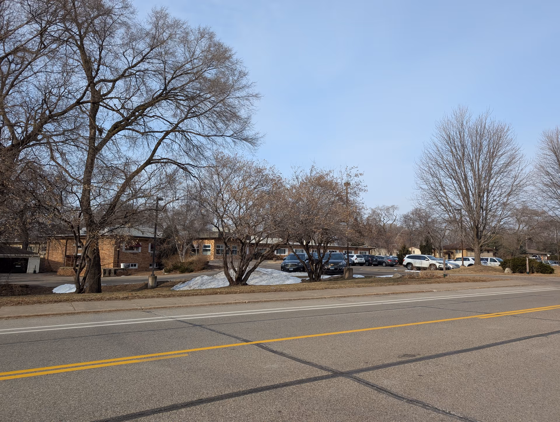 Low single-story brick senior living buildings and a parking lot behind leafless trees with a road in the foreground.