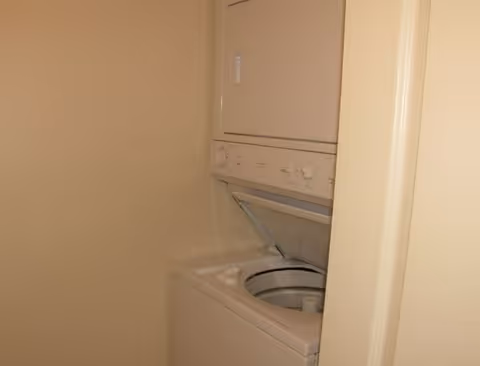 A small laundry area with a stacked washer and dryer unit. The washer lid is open, showing the inside of the washing machine. The walls around the unit are plain and light-colored.