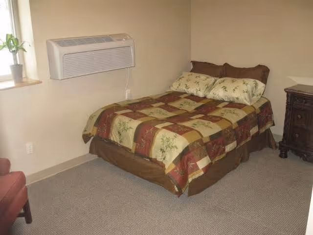 Small furnished bedroom with a double bed covered by a patterned quilt, a wooden bedside dresser, wall-mounted air conditioner, and a potted plant on the windowsill.