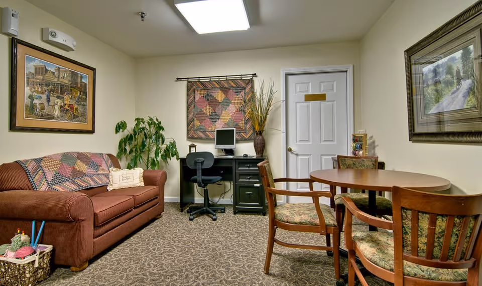 A cozy senior living common area featuring a brown couch with a colorful quilt draped over it, a small desk with a computer and office chair, a round wooden table with four cushioned chairs, framed artwork on the walls, a potted plant, and a basket with yarn and knitting needles on the floor.