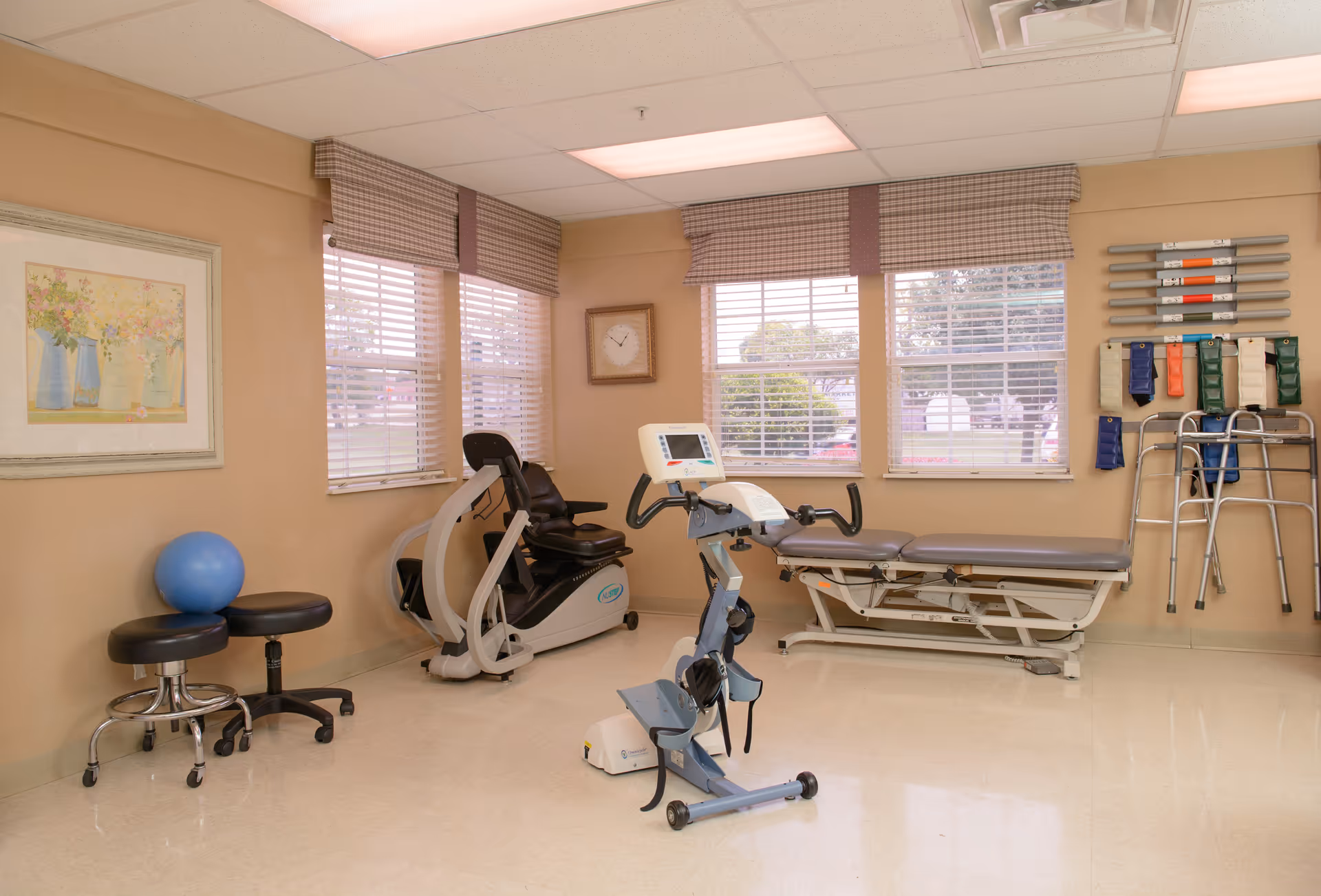 A physical therapy or rehabilitation room with exercise equipment including a recumbent bike, a stationary bike, and a therapy table. There are two stools, a blue exercise ball, and various resistance bands and walkers hanging on the wall. The room has beige walls, three windows with blinds and valances, a framed floral painting, and a clock on the wall.