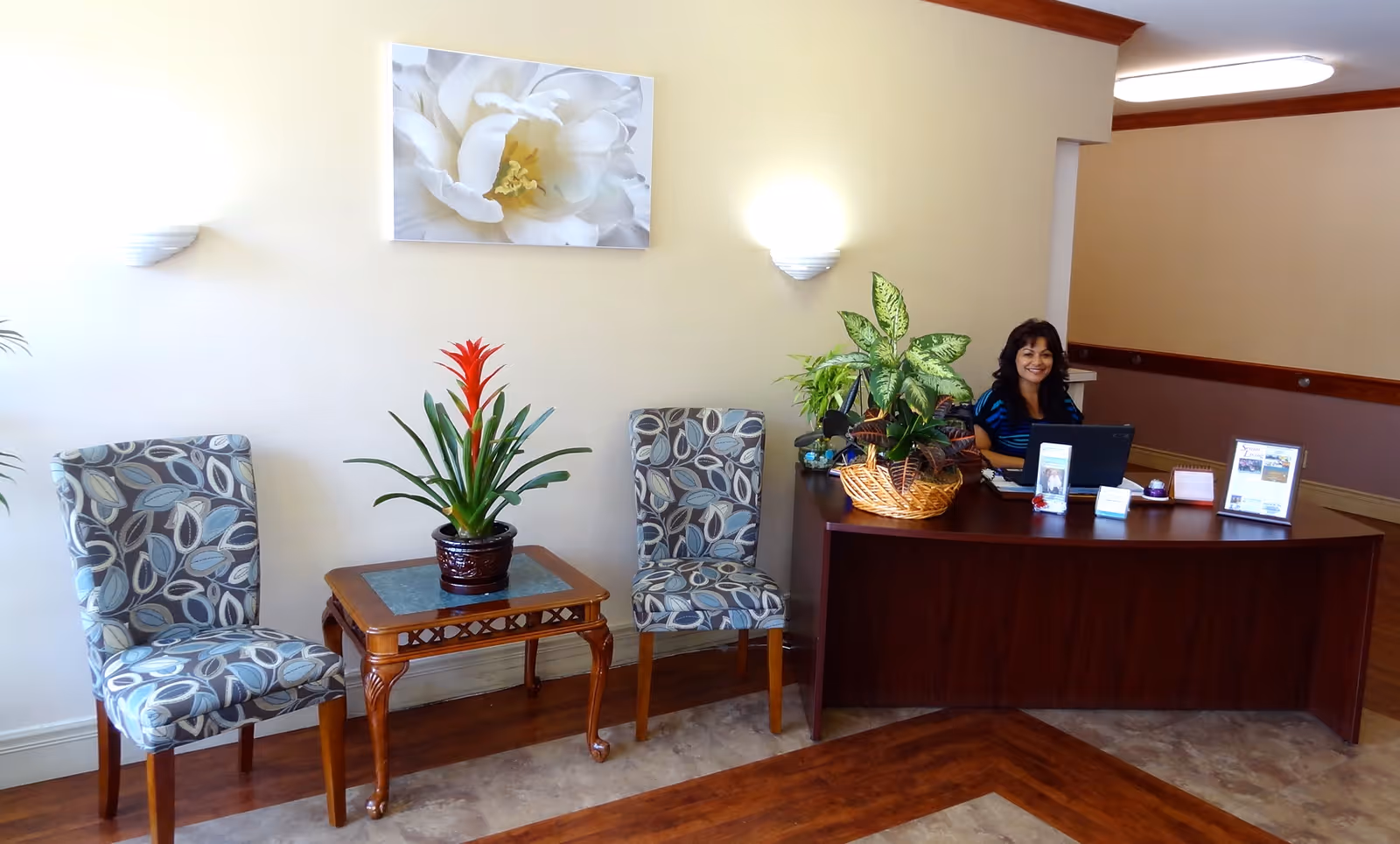 Reception area with two patterned chairs and a small table with a potted plant, and a smiling receptionist behind a wooden desk with plants and a laptop.
