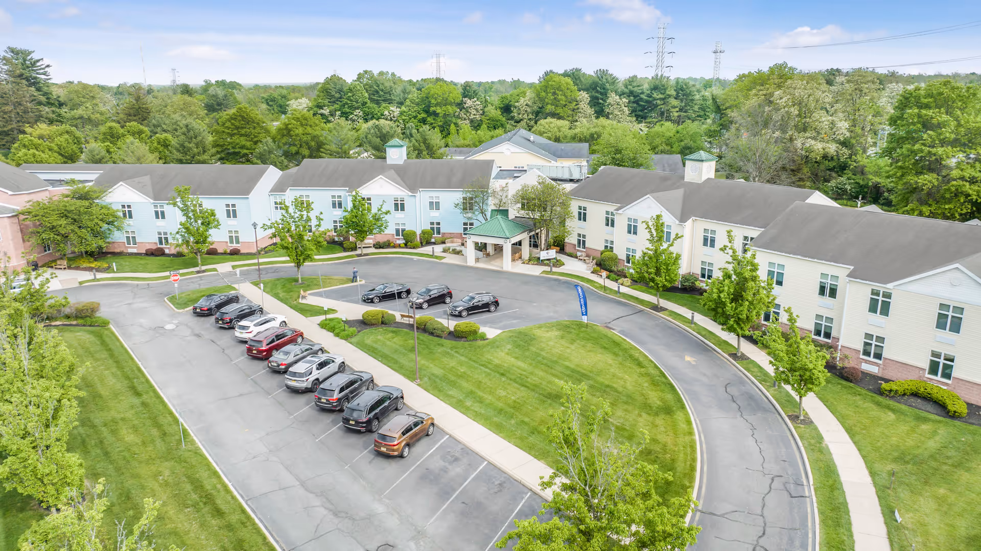 Aerial view of The Terraces at Lawrence senior living facility showing a large two-story building with a green roofed entrance, surrounded by well-maintained lawns, trees, and a parking lot with several cars parked.
