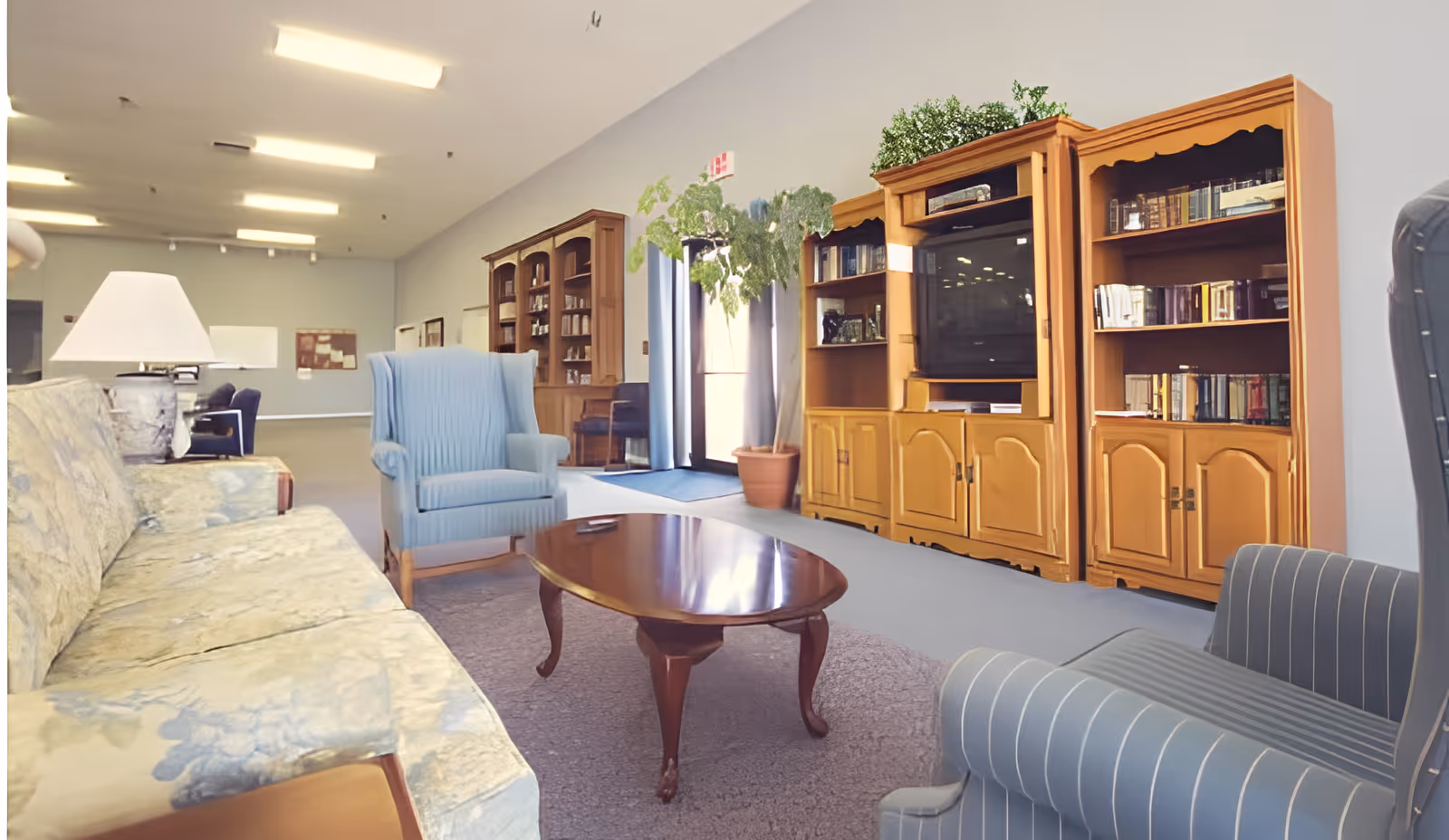 A spacious living room area in an assisted living facility featuring a floral patterned sofa, two blue upholstered armchairs, a wooden coffee table, and a large wooden entertainment center with bookshelves and a television. There is a potted plant near the entrance door with glass panels, and the room is well-lit with ceiling lights.