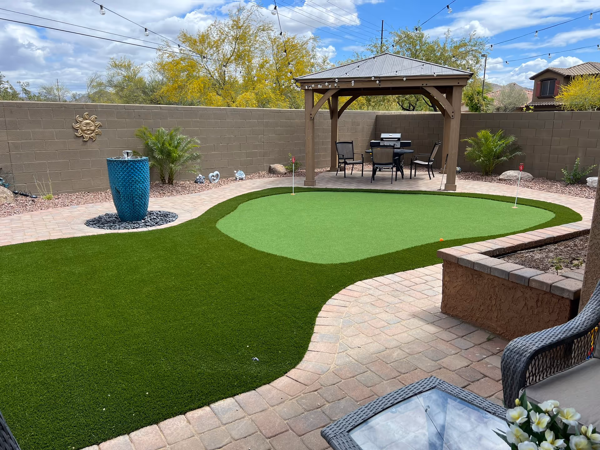 Backyard patio featuring an artificial putting green, a gazebo with seating and grill, a blue decorative fountain, and string lights.