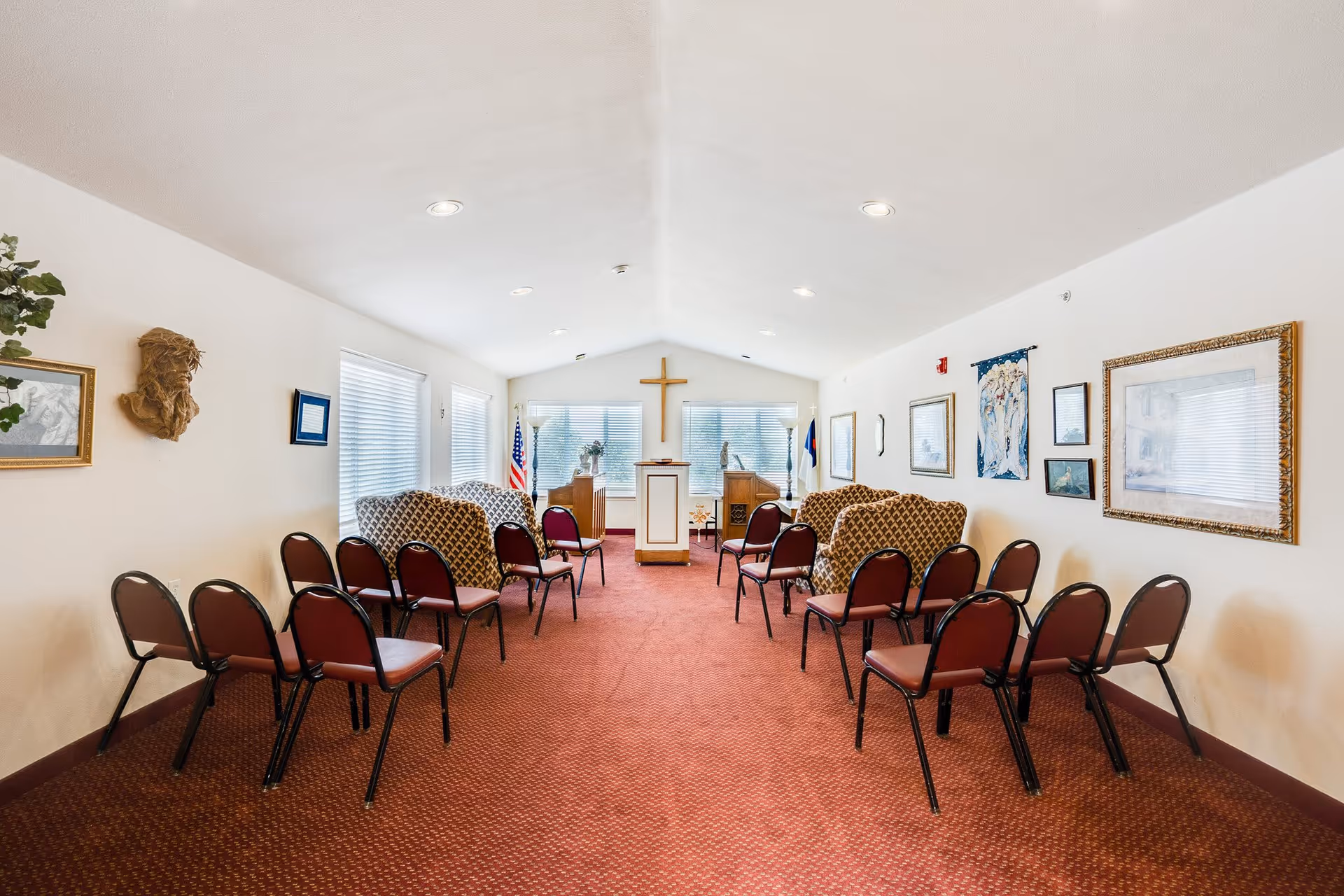 Interior view of a small chapel or worship room with rows of chairs facing a podium and a cross mounted on the far wall. The room has carpeted flooring, white walls adorned with framed pictures and flags, and ceiling lights.