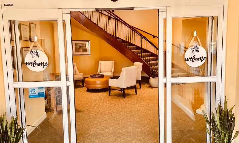 Entrance view through glass doors into a cozy seating area with four beige upholstered chairs arranged around a round brown ottoman. A staircase with wooden railing is visible in the background. Two round welcome signs hang on the glass doors. Potted plants are placed near the entrance.