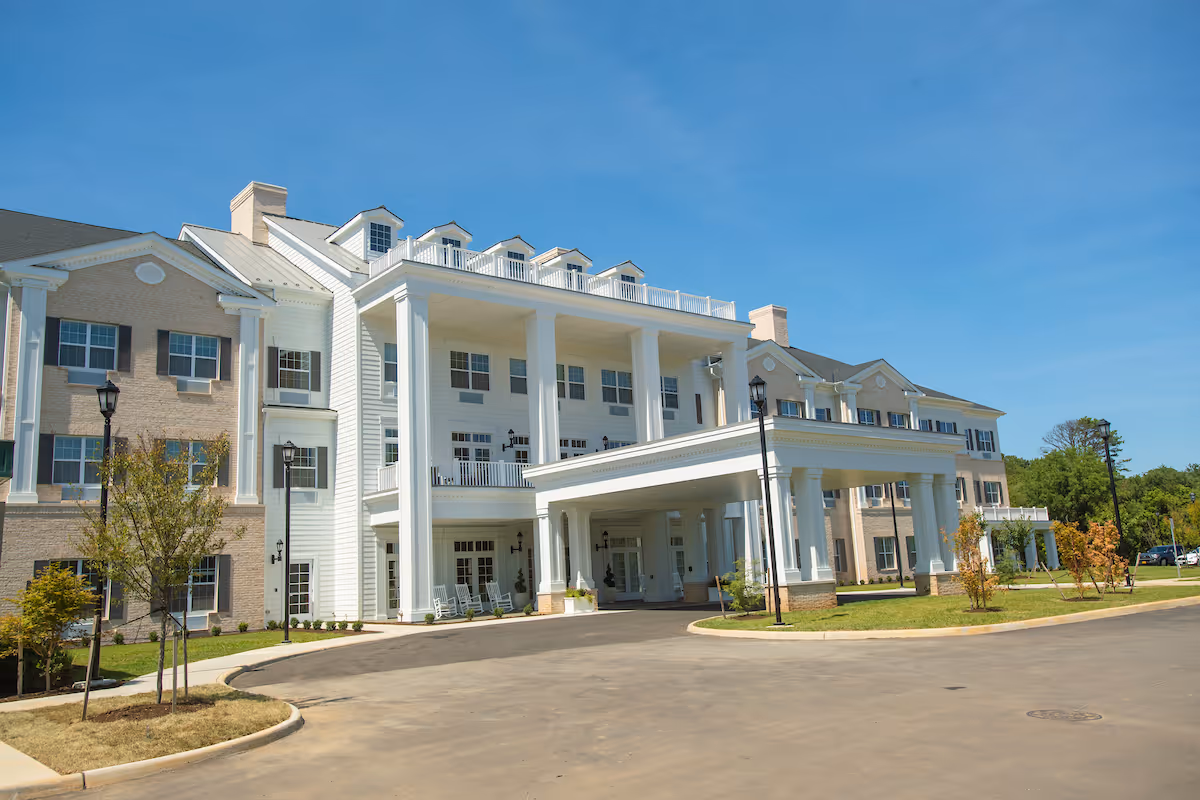 Exterior view of a large, elegant senior living facility building with white columns and a covered entrance. The building has multiple windows, a mix of brick and white siding, and a clear blue sky in the background. There are small trees and landscaped areas around the driveway.