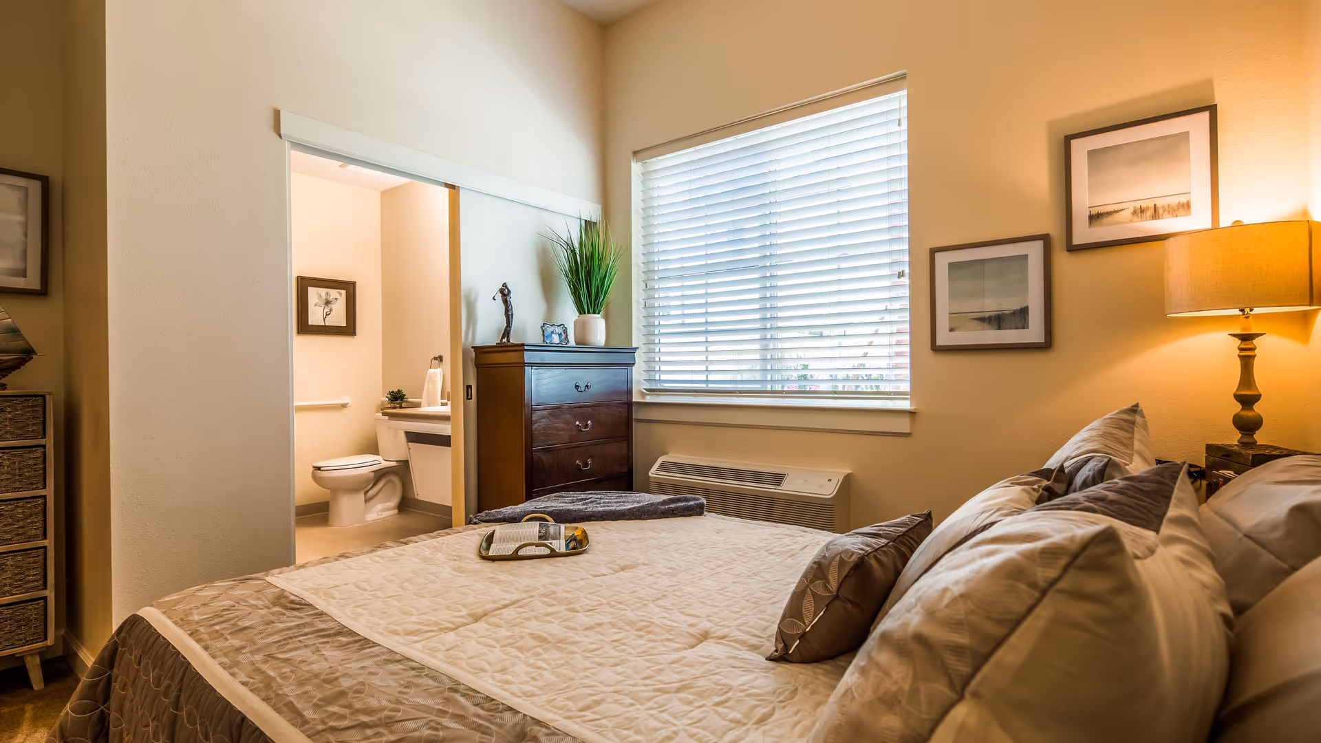 A cozy bedroom with a neatly made bed featuring multiple pillows and a beige quilt. There is a wooden dresser with decorative items and a potted plant next to a window with white blinds. On the wall above a bedside table with a lamp are two framed pictures. An open door reveals a bathroom with a toilet, sink, and framed artwork on the wall.