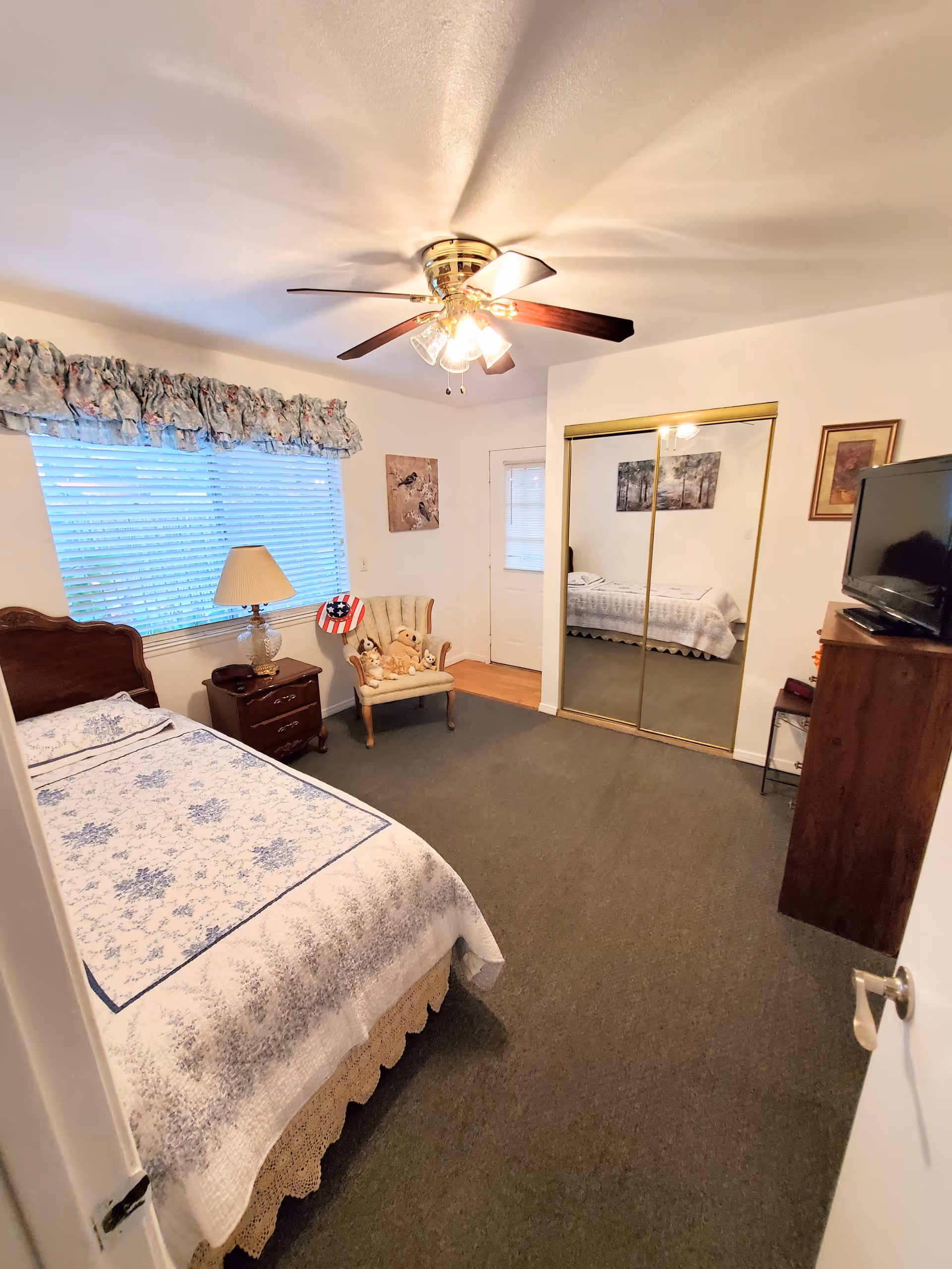 Bedroom with a single bed covered in a patterned quilt, nightstand and lamp, armchair with stuffed animals, mirrored closet doors, a dresser with TV, and a ceiling fan.