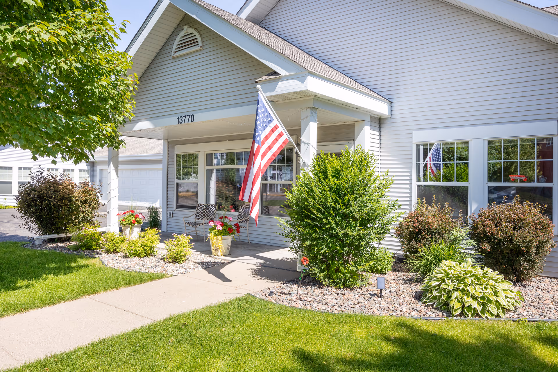 Front exterior view of a senior living facility named The Farmstead with a covered entrance, American flag, two chairs, potted flowers, and well-maintained landscaping including bushes, a tree, and green grass.
