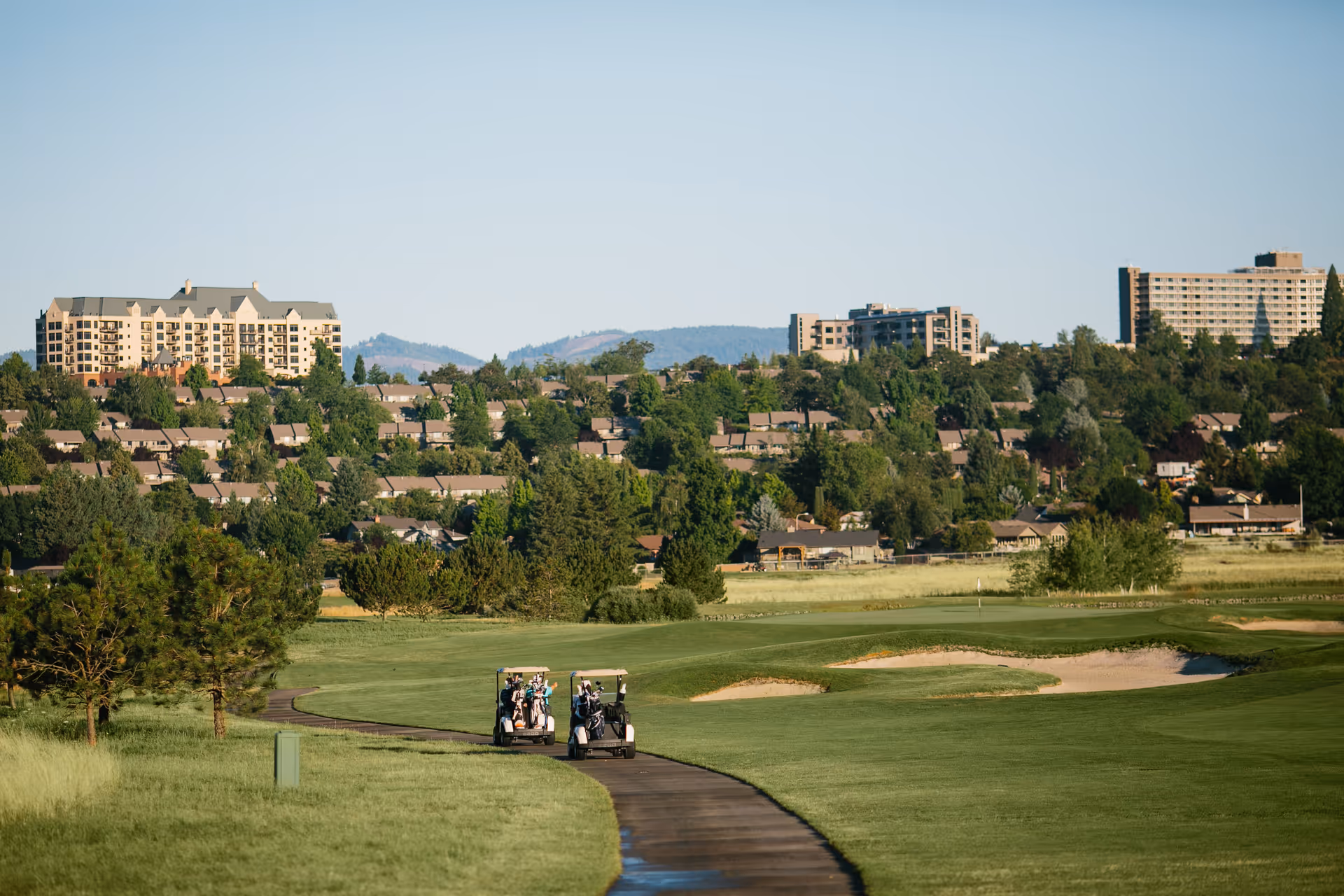 Golf course fairway with two golf carts on a paved path and residential buildings on a tree-covered hillside in the background.
