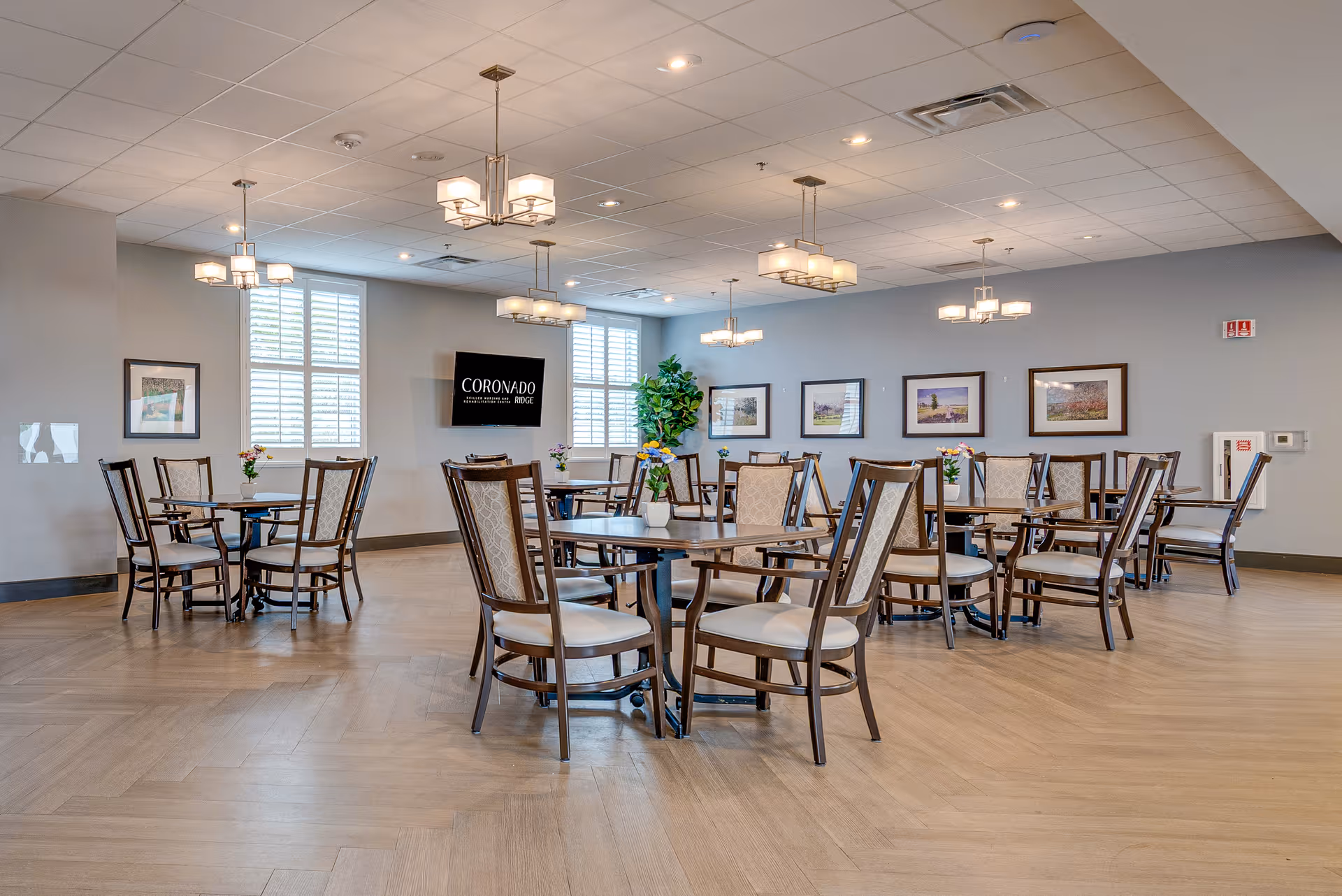 Bright dining room with round tables and cushioned chairs, modern chandeliers, framed artwork, and a wall-mounted TV reading "Coronado".