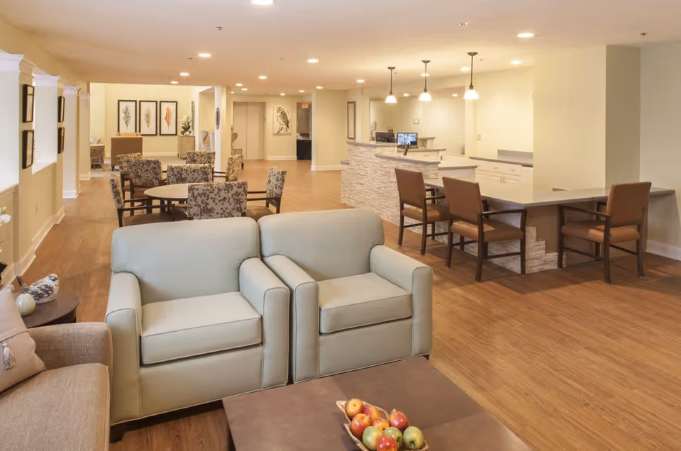 A spacious and well-lit common area in a senior living facility featuring comfortable seating including two light blue armchairs and a beige sofa around a coffee table with a bowl of fruit. In the background, there are multiple tables and chairs, a reception desk with pendant lights hanging above, and framed artwork on the walls. The flooring is wood, and the walls are painted a soft yellow.
