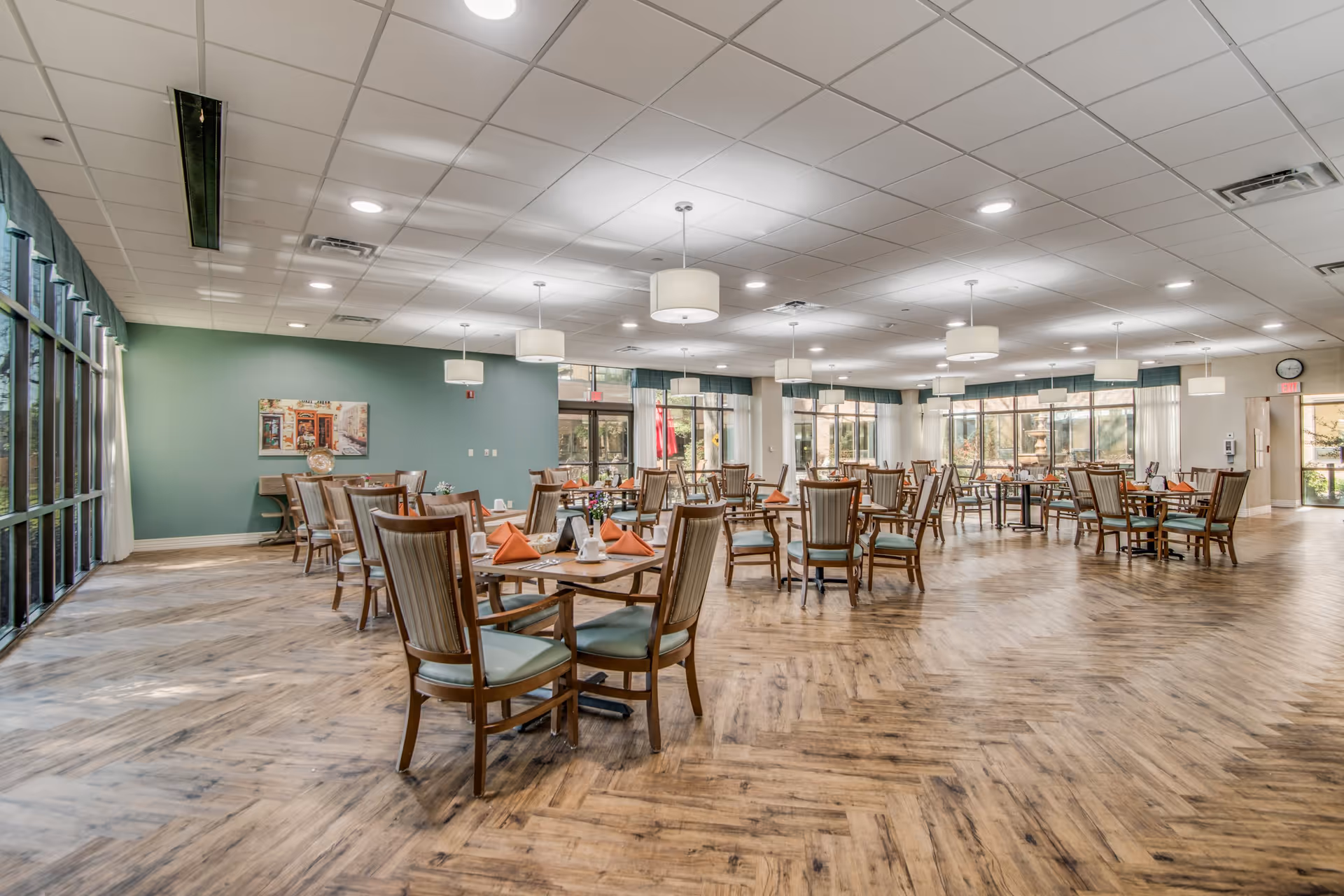 Spacious dining room with multiple wooden tables and chairs arranged neatly. Each table is set with orange napkins, white cups, and small flower arrangements. Large windows on two sides allow natural light to fill the room, and modern ceiling lights hang above the tables. The floor has a wood pattern, and a green accent wall features a framed picture.
