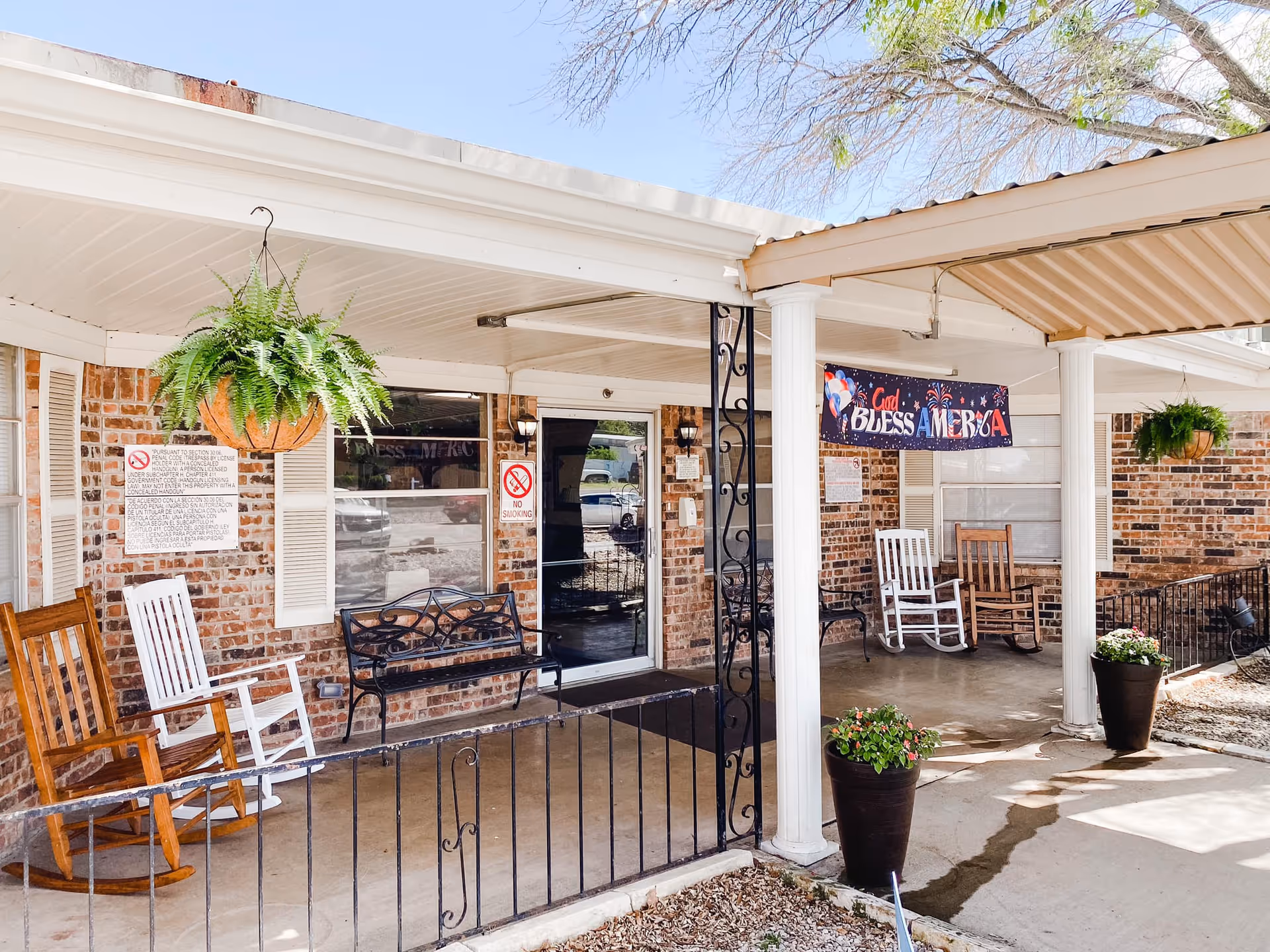 Covered front porch of a brick nursing home with rocking chairs, hanging ferns, potted plants, and a 'God Bless America' banner.