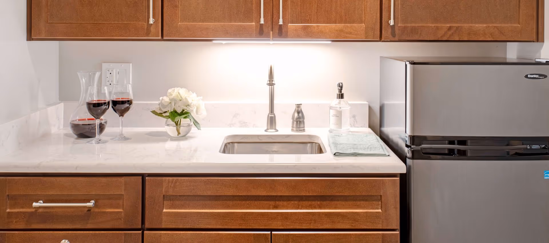 A kitchen countertop with a stainless steel sink, a modern faucet, and wooden cabinets above and below. On the countertop, there is a glass carafe and two wine glasses filled with red wine, a small vase with white flowers, a soap dispenser, and a folded green towel. A stainless steel mini refrigerator is positioned to the right of the countertop.