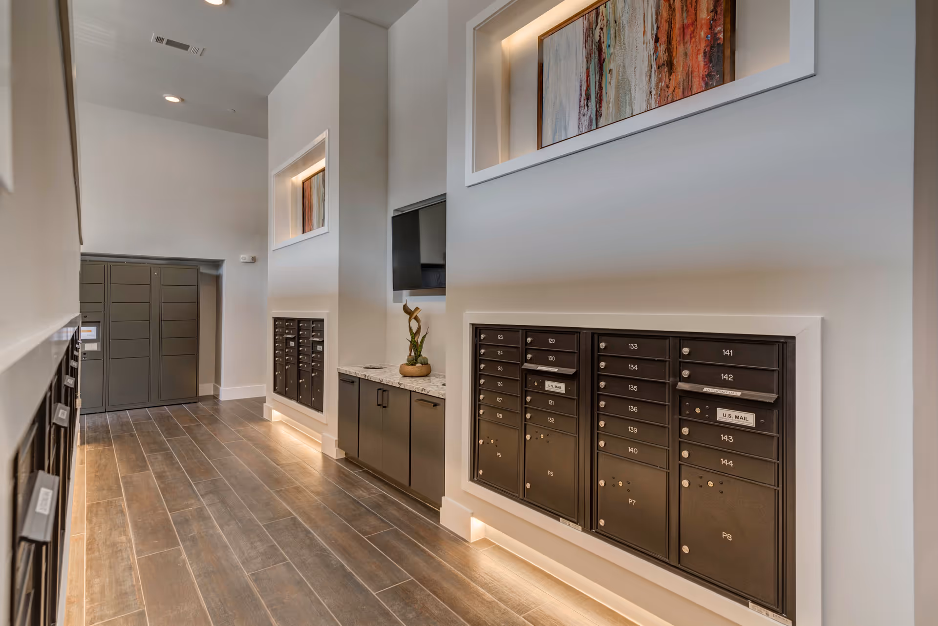 A modern interior mailroom hallway with wall-mounted resident mailboxes, parcel lockers, and cabinetry beneath recessed artwork.
