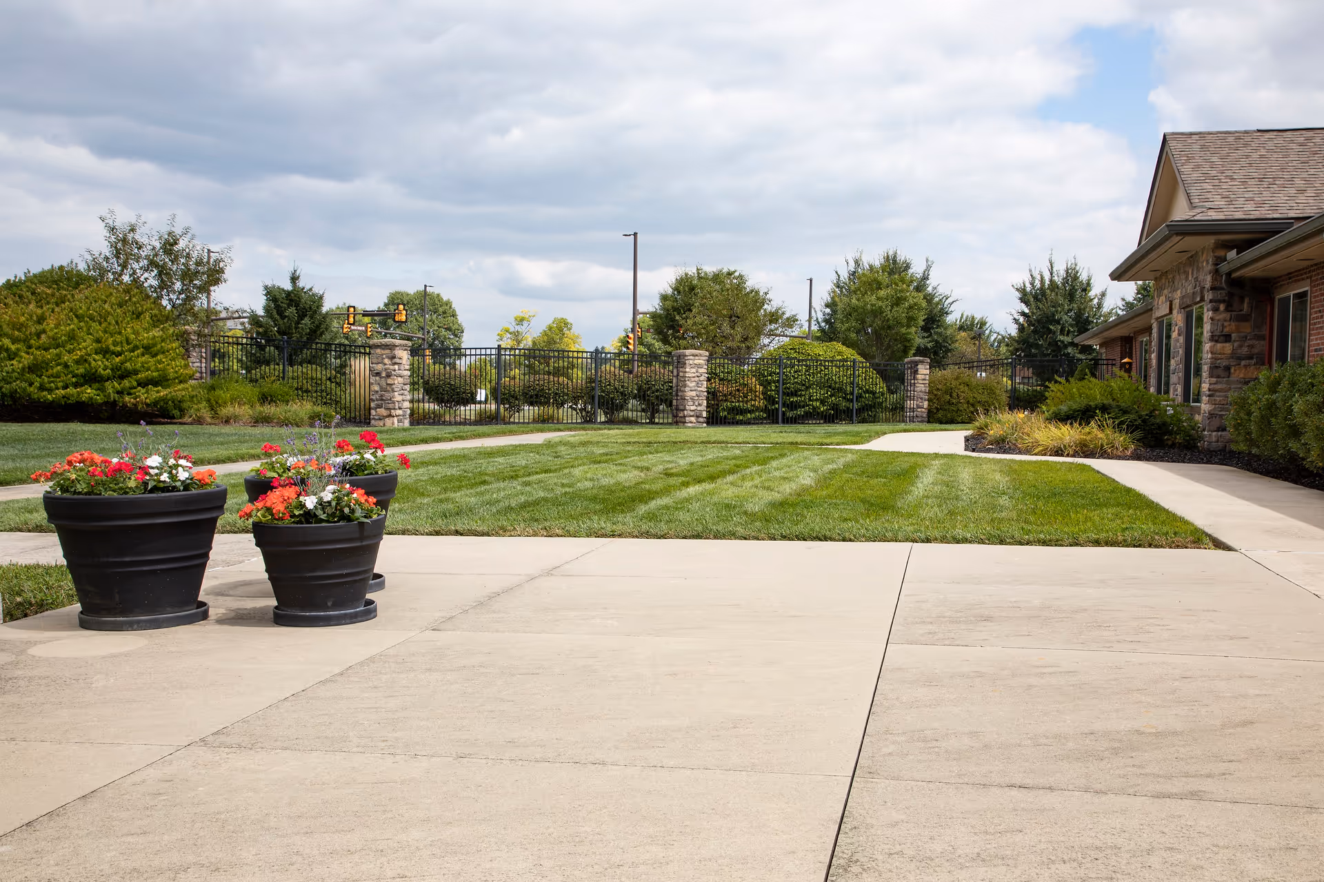 Outdoor area of Dublin Glenn Memory Care featuring a concrete patio with three black flower pots containing red and white flowers, a well-maintained green lawn, a paved walkway, and a building with stone and brick exterior on the right. The background includes a black metal fence with stone pillars, trees, and a partly cloudy sky.