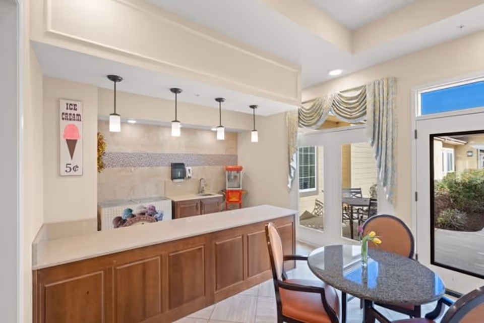 Interior view of a small dining and snack area in a memory care community. The space features a counter with wooden cabinets, a popcorn machine, a sink, and a wall-mounted paper towel dispenser. Above the counter, there are four hanging pendant lights. To the right, there is a round table with two chairs and a small vase with yellow flowers. Large windows and a glass door provide a view of an outdoor patio area with seating. A decorative curtain hangs above the windows and door. On the left wall, there is a vintage-style sign advertising ice cream for 5 cents.