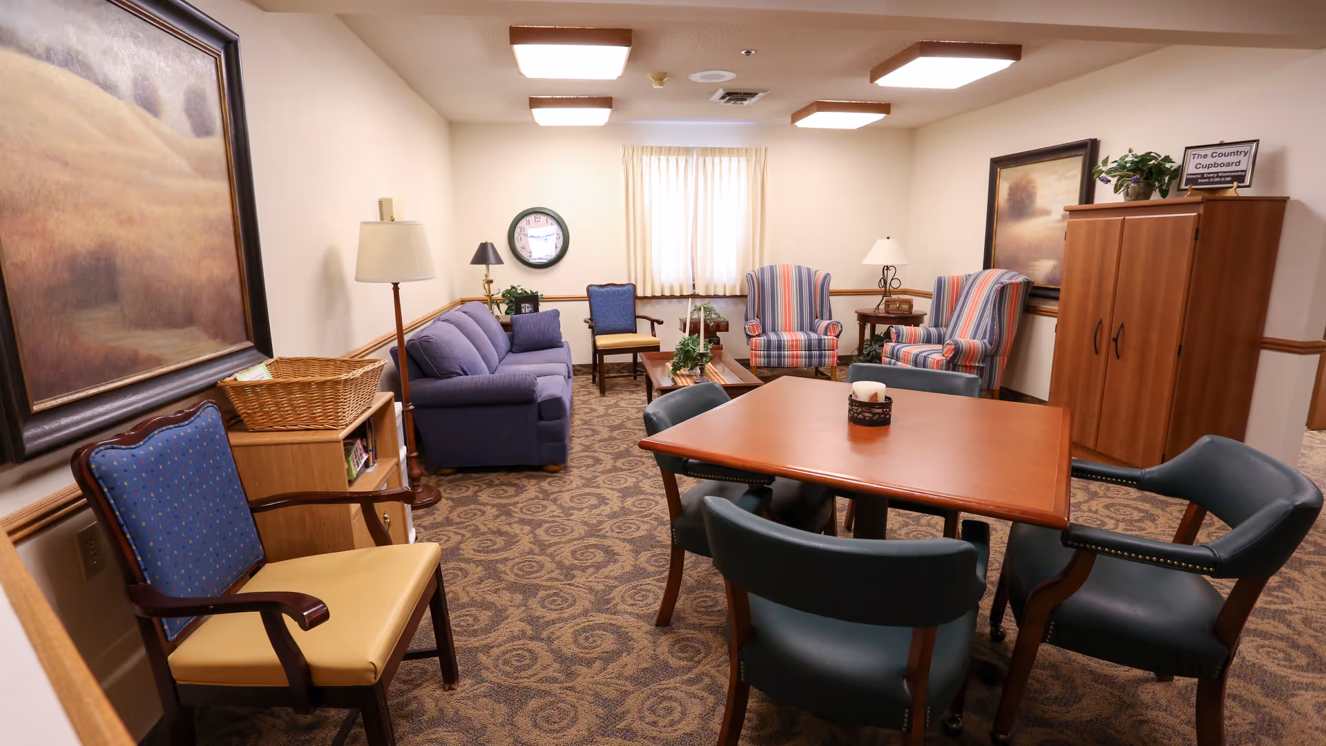 Community lounge with a central table and chairs, sofas and armchairs, lamps and artwork in a senior living facility.
