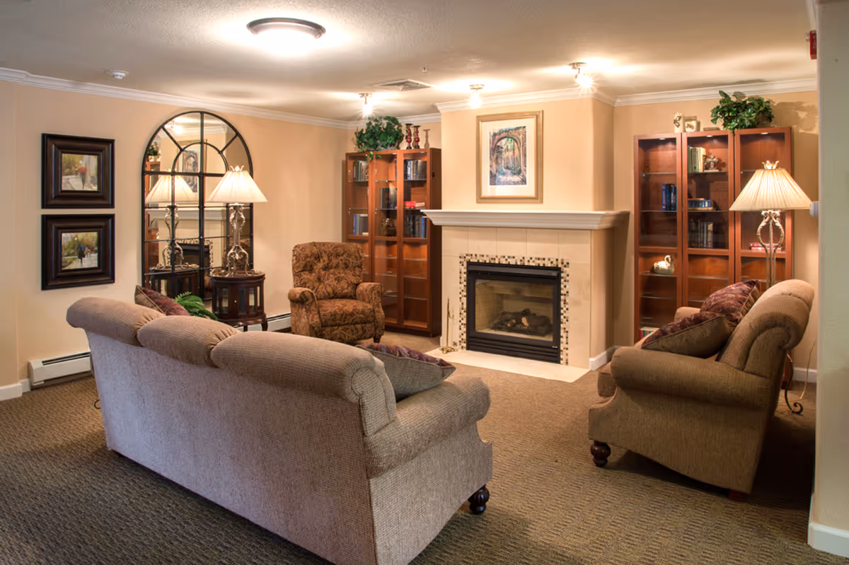 A cozy living room with two beige sofas and a patterned armchair arranged around a fireplace. The fireplace has a framed picture above it and is flanked by two wooden cabinets with glass doors displaying books and decorative items. There are two table lamps with beige shades on either side of the room, a large decorative mirror on one wall, and two framed paintings. The room has beige walls and carpeted flooring.